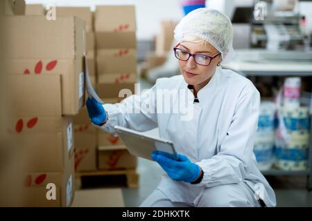 Young beautiful focused female worker in sterile cloths using a tablet to check correction of inventory in factory storage room. Stock Photo