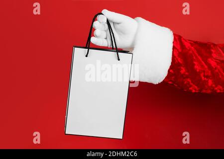 Santa hand with white shopping bag on red background. Banner, flyer or poster mock up for Christmas sale. Paper bag for shopping  Stock Photo