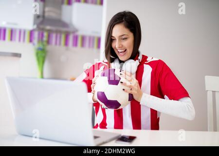 Young beautiful happy girl in red and white jersey is watching a soccer match cheering on her laptop and holding a ball with both hands. Stock Photo