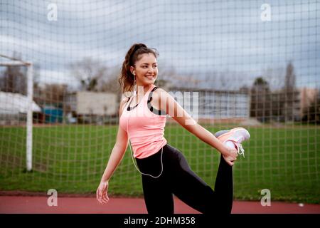 Beautiful young fitness girl doing leg stretching while standing on one leg near the soccer field. Stock Photo