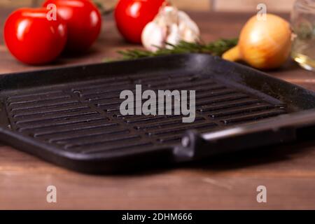 empty grill pan, next to tomatoes, onions and garlic on a wooden table. Stock Photo