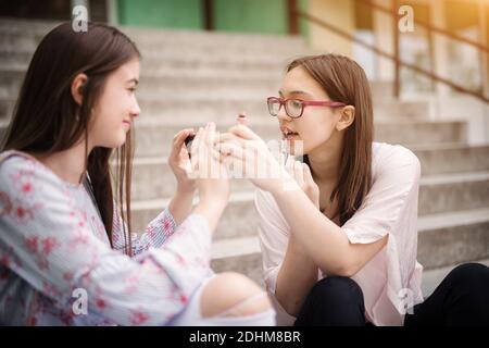 Two adorable charming high school girls applying makeup while sitting on stairs outside the school.