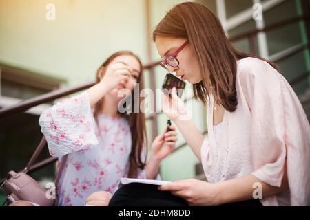Hardworking smart high school girl reading school notes and sitting on stairs outside the school while another girl applying makeup.