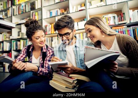 A group of young focused joyful students studying, explaining each other and writing notes in a notebook while sitting on a floor in front of a librar Stock Photo