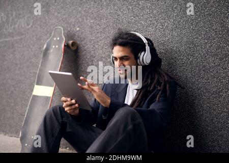 Young cheerful stylish man with dreadlocks is sitting and leaning against the wall with his longboard and listening to music with on tablet. Stock Photo