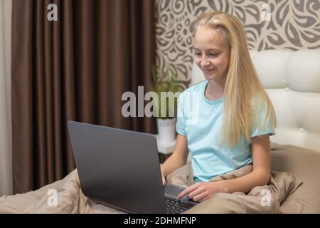 A teenage girl with blond hair works on a laptop in bed. Distance education theme. high quality Stock Photo