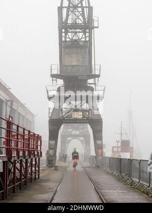 A cyclist rides along the quayside of Bristol's Floating Harbour where old dock cranes are shrouded in fog outside the M Shed Museum. Stock Photo