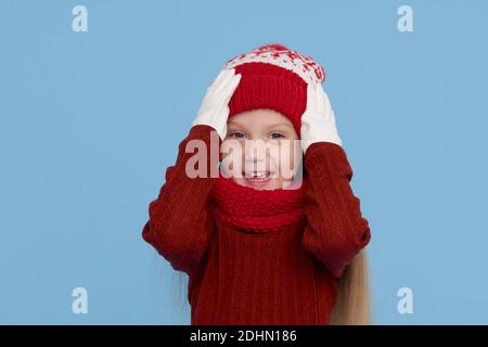 Winter portrait of happy child girl wearing knitted red hat, snood and sweater. Wow face. Blue background. childrens winter entertainment Stock Photo