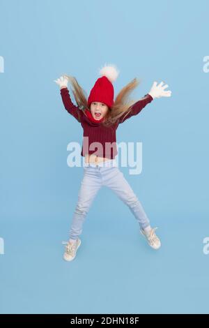 Winter portrait of happy child girl in a knitted red hat, snood and sweater jumping high for joy. Blue background. childrens winter entertainment Stock Photo