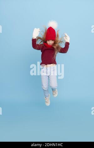Winter portrait of happy child girl in a knitted red hat, snood and sweater jumping high for joy. Blue background. childrens winter entertainment Stock Photo