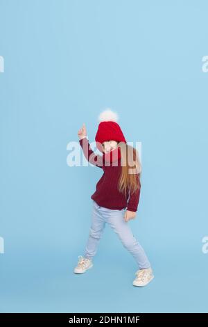 Winter portrait of happy child girl in a knitted red hat, snood and sweater points up. Blue background. childrens winter entertainment Stock Photo