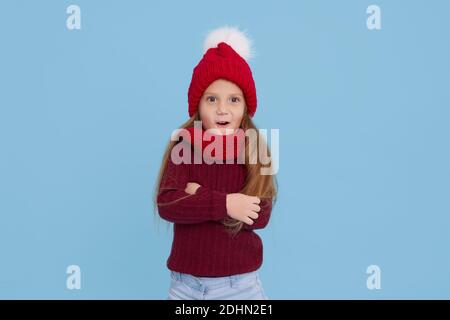 Winter portrait of happy child girl wearing knitted red hat, snood and sweater. Wow face. Blue background. childrens winter entertainment Stock Photo