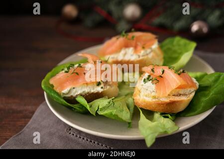 Baguette canapes with salmon and cream cheese on a plate with lettuce, dark rustic wooden table with Christmas decorations, copy space, selected focus Stock Photo