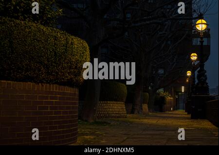 Nighttime on a pathway along red brick wall, hedges and street lamps leaving yellow glow Stock Photo