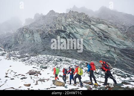 Group of male alpinists with backpacks and trekking sticks having winter hiking tour in mountains, walking on rocky path covered with snow. Concept of travelling, hiking and mountaineering. Stock Photo
