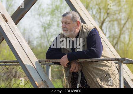 Portrait of Caucasian senior man with walking stick standing on an metal pedestrian bridge and looking at the distance Stock Photo
