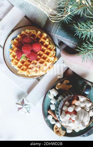Viennese waffles with raspberries and honey on the Christmas table with a hot drink with marshmallows and Christmas tree branches with garlands of lig Stock Photo