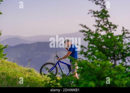Tired cyclist is wiping his sweat off his face while pushing his bicycle uphill on a dirt road in a mountain. Stock Photo