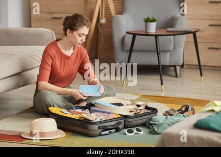 Young woman packing masks in suitcase while sitting on the floor and preparing for her vacation Stock Photo
