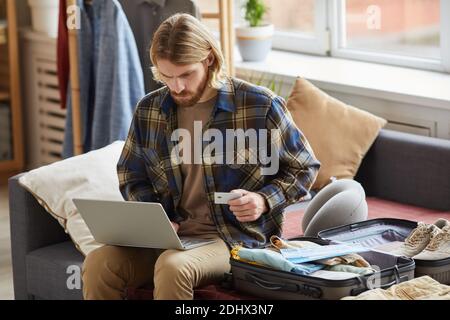 Young man using laptop and credit card for online payment while sitting on sofa with luggage Stock Photo