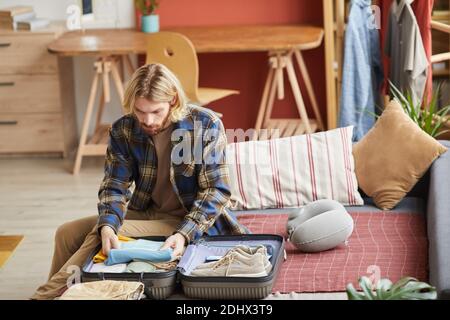 Young man sitting in the room packing his clothes in suitcase he is going in business trip Stock Photo
