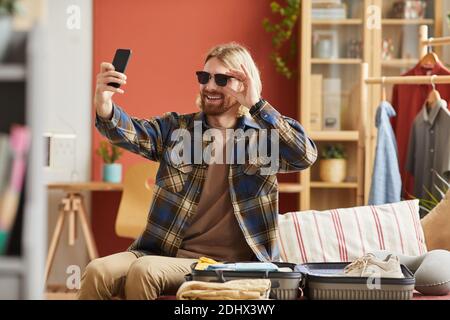 Young happy man wearing sunglasses making selfie on mobile phone while packing clothes at home Stock Photo