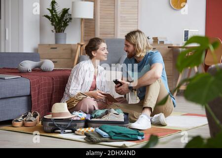 Young couple packing their clothes in suitcase sitting on the floor in the room and talking Stock Photo
