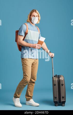 Portrait of young tourist in protective mask standing with luggage against the blue background Stock Photo