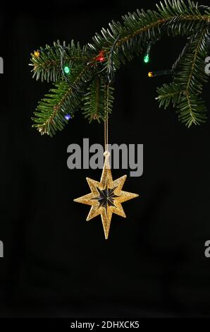 Closeup of Christmas tree with lights and hanging gold glitter star against a black background Stock Photo