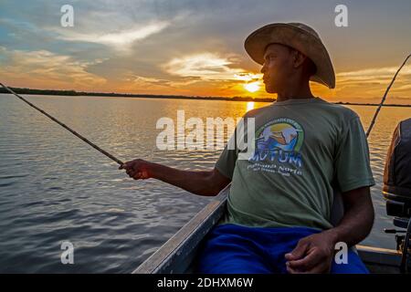 At sunset,  a Brazilian angler relaxing on the back of a boat. using a bamboo cane as a fishing rod, waiting for a bite from a Yellow belly Piranha in Stock Photo