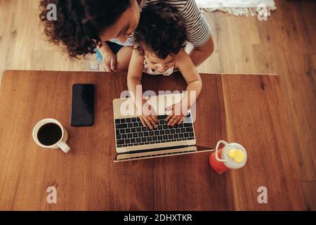 Top view of a woman watching her baby using laptop. Child sitting on mother's lap and using laptop at table. Stock Photo