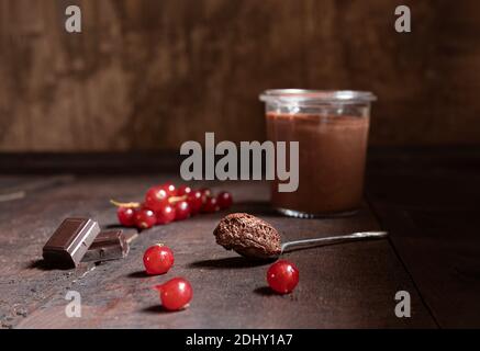 A glass and a teaspoon with chocolate mousse, pieces of dark chocolate and red currant berries on the wooden background Stock Photo