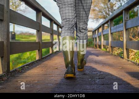 Low angle, rear view of UK woman in wellington boots/ wellies, isolated outdoors walking in winter sunshine crossing over a wooden footbridge. Stock Photo