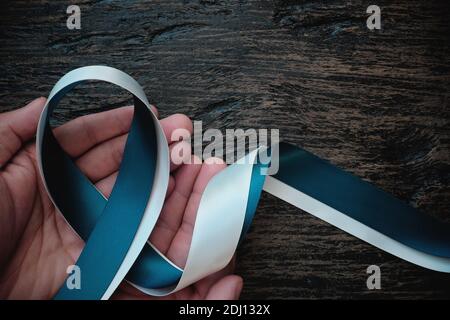 Top view of hand holding white and teal ribbon on dark wooden background with copy space. Cervical cancer awareness month, care and hope concept. Stock Photo