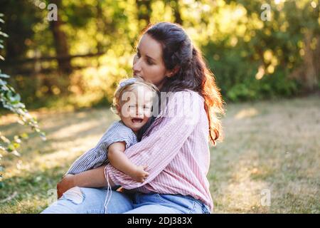 Mother hugging pacifying sad upset crying toddler girl. Family young mom and crying baby in park outdoor. Bonding relationship of mom and child baby. Stock Photo