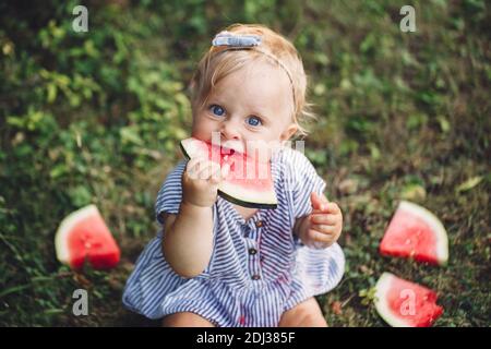 Cute Caucasian baby girl eating watermelon in park. Funny child kid sitting on ground with fresh fruit outdoor. Supplementary healthy finger food for Stock Photo