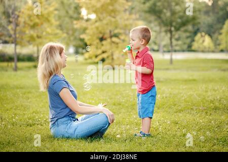 Mothers Day. Mother and boy toddler son blowing soap bubbles in park. Mom and child playing having fun together outdoor on summer day. Happy authentic Stock Photo