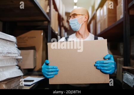 Tired warehouse worker carrying box, wearing mask and gloves. Selective focus Stock Photo