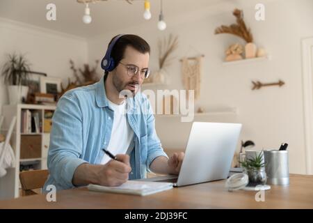 Focused man wearing headphones writing notes, looking at laptop screen Stock Photo