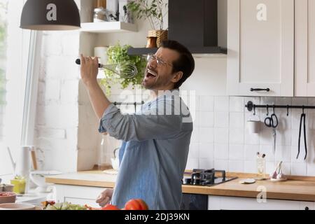 Funny man wearing glasses singing into whisk in kitchen Stock Photo