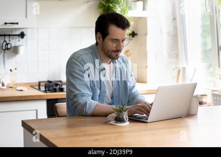 Confident young man wearing glasses using laptop, working in kitchen Stock Photo