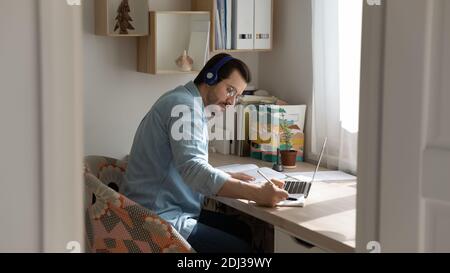Focused man wearing headphones studying online, writing notes, using laptop Stock Photo