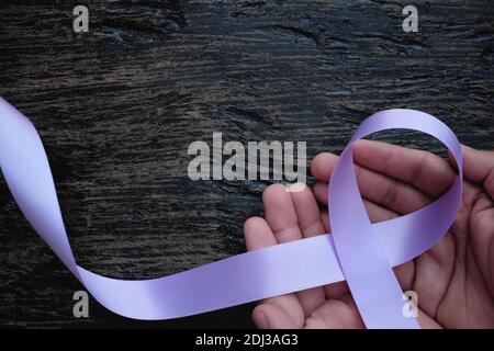 Top view of hands holding lavender or light purple orchid color ribbon on dark wooden background. General and testicular cancer and epilepsy awareness Stock Photo