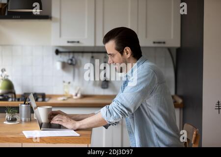 Smiling man wearing glasses working on laptop in modern kitchen Stock Photo