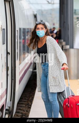 Young woman using a surgical mask holding two suitcases next to to a train while looking at the camera. Tourism and safety concept. Stock Photo