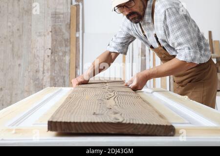 Male carpenter working the wood in carpentry workshop, holding wooden plank, wearing overall Stock Photo