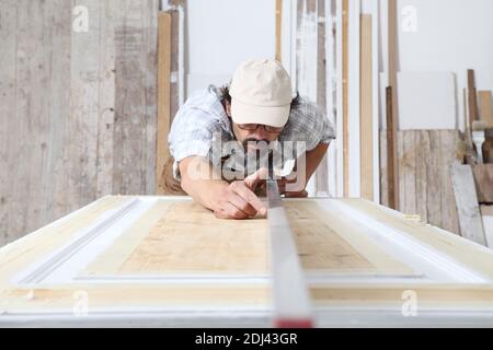 Male carpenter working the wood in carpentry workshop, checking level of wooden door with bubble level, wearing overall Stock Photo