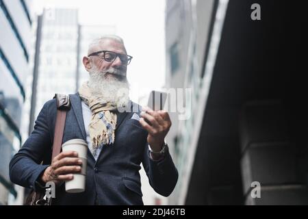 Business hipster senior man using mobile phone and drinking coffee with city in background - Focus on face