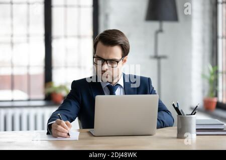 Confident businessman wearing glasses using laptop, writing notes Stock Photo