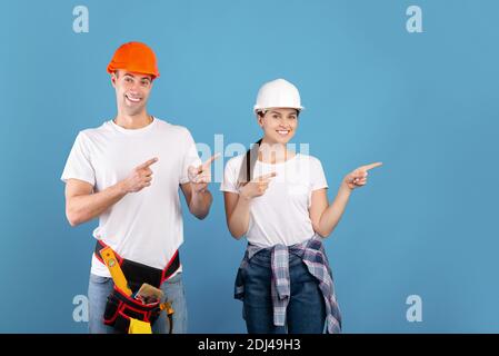 Smiling handyworkers couple wearing casual clothes and protective hard hats pointing aside Stock Photo
